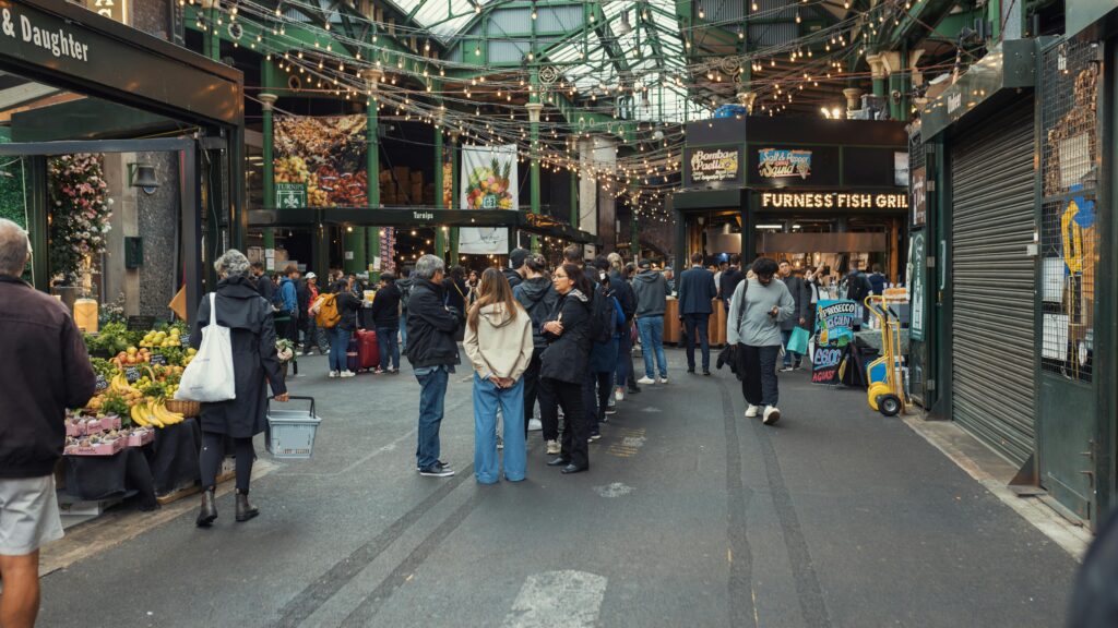 A bustling indoor market scene with diverse shoppers exploring food stalls under decorative lights.