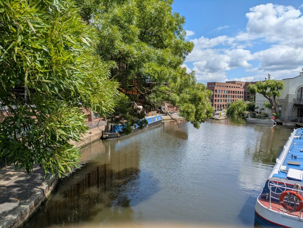 Scenic view of a tranquil canal with moored boats and lush greenery in London under a clear blue sky.