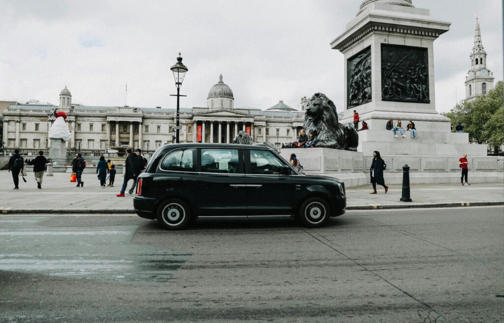 A classic black taxi cab parked in front of London's iconic Trafalgar Square with people and historic architecture.